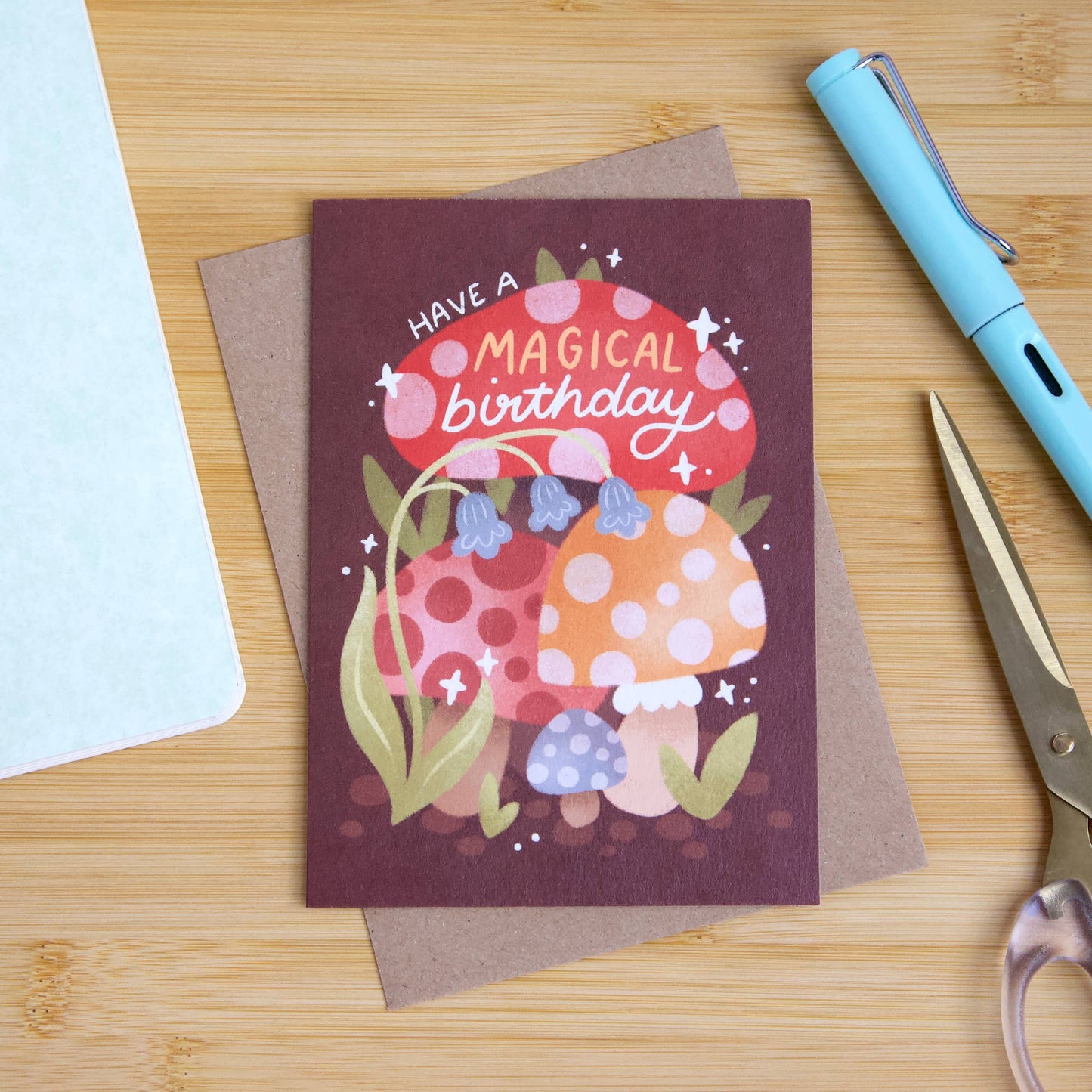 Greeting card with mushrooms and 'Have a Magical Birthday' text on a wooden surface with stationery items.