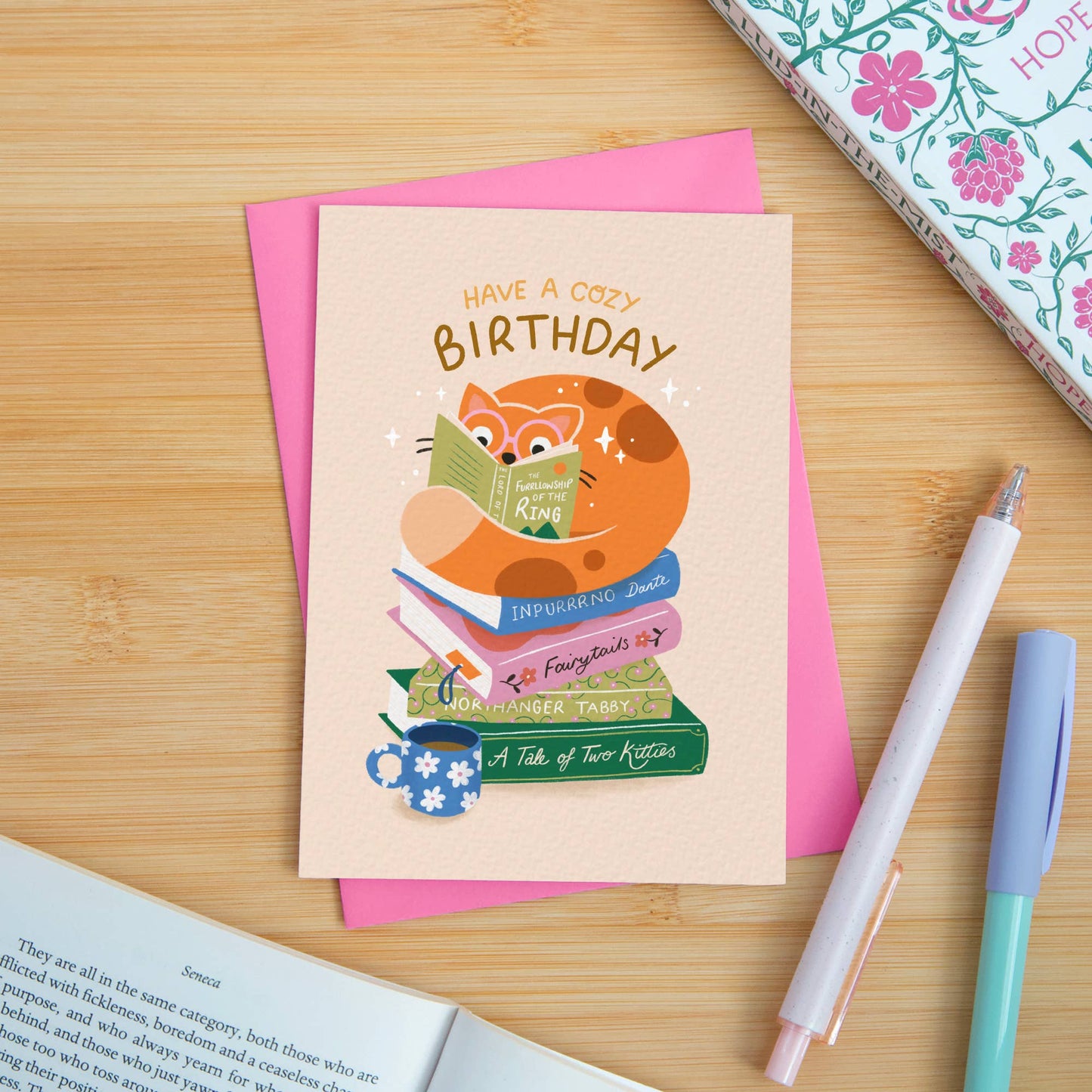 Birthday card with fox reading a book on a wooden surface with pens and books.