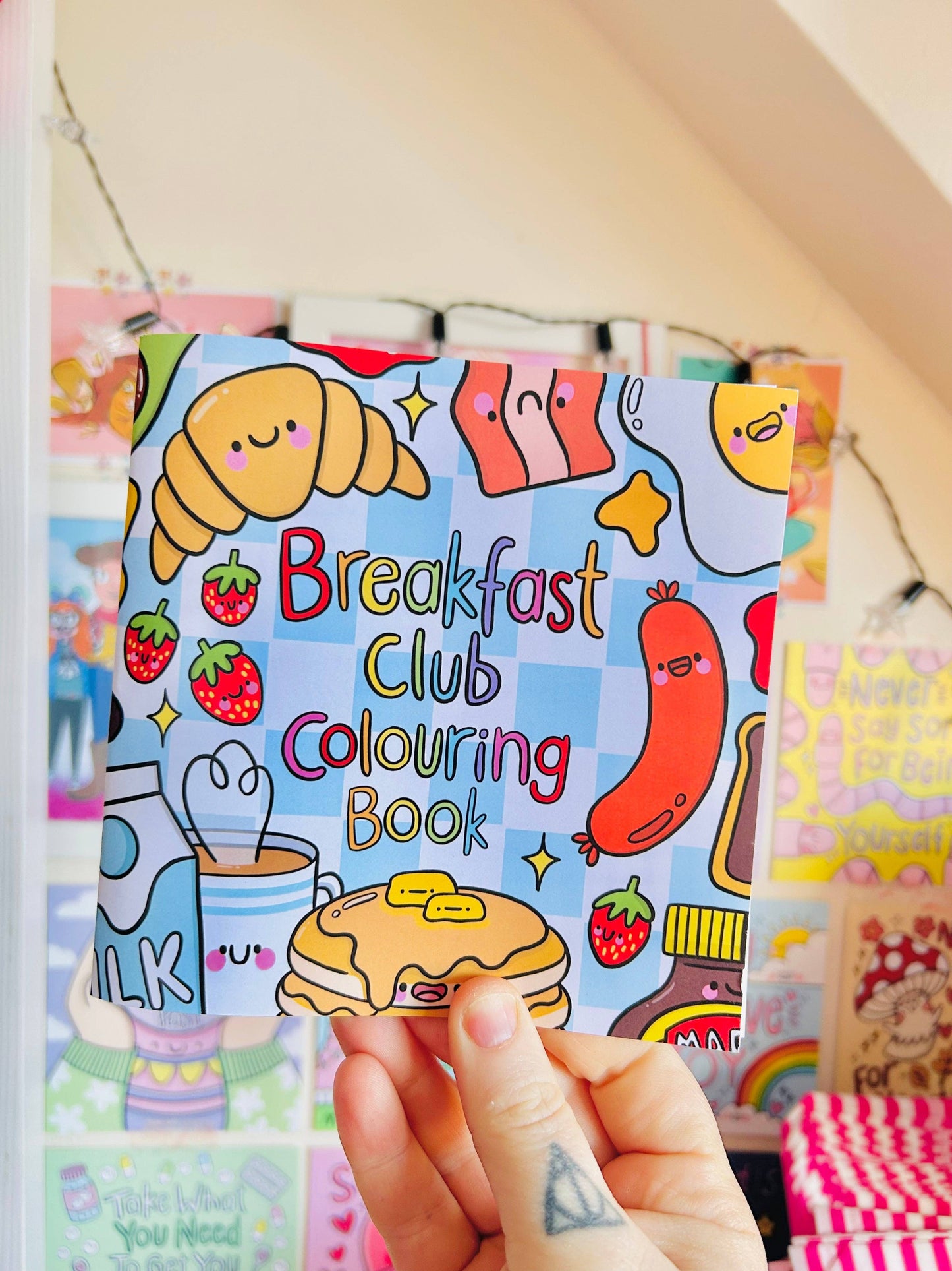 Hand holding a 'Breakfast Club Colouring Book' with colorful illustrations of food.
