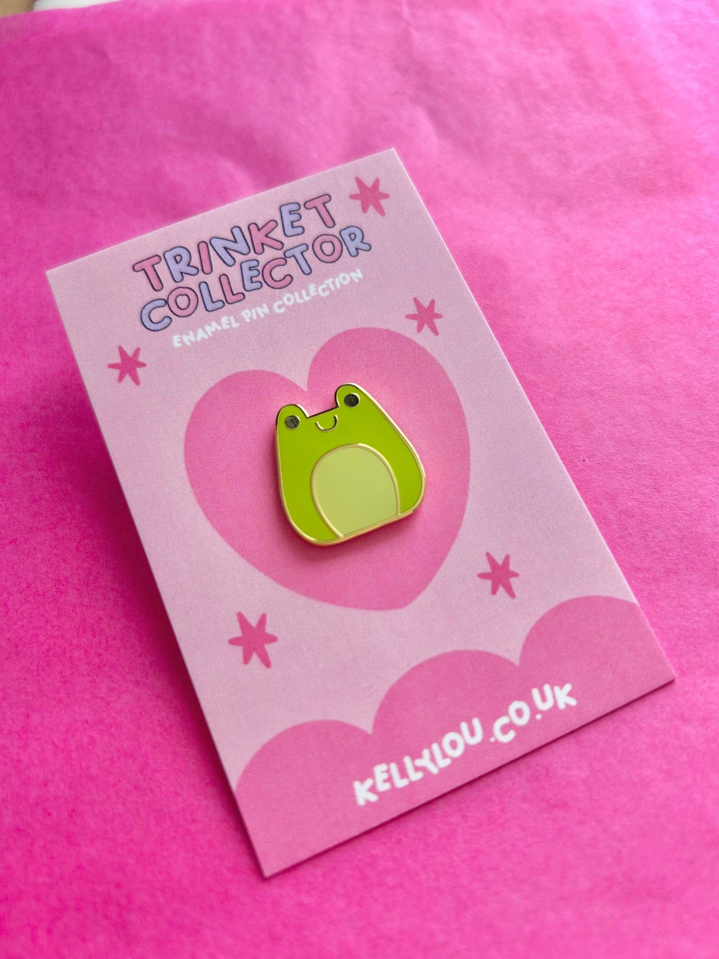 Green frog-shaped pin on a pink card with 'Trinket Collector' branding against a pink background