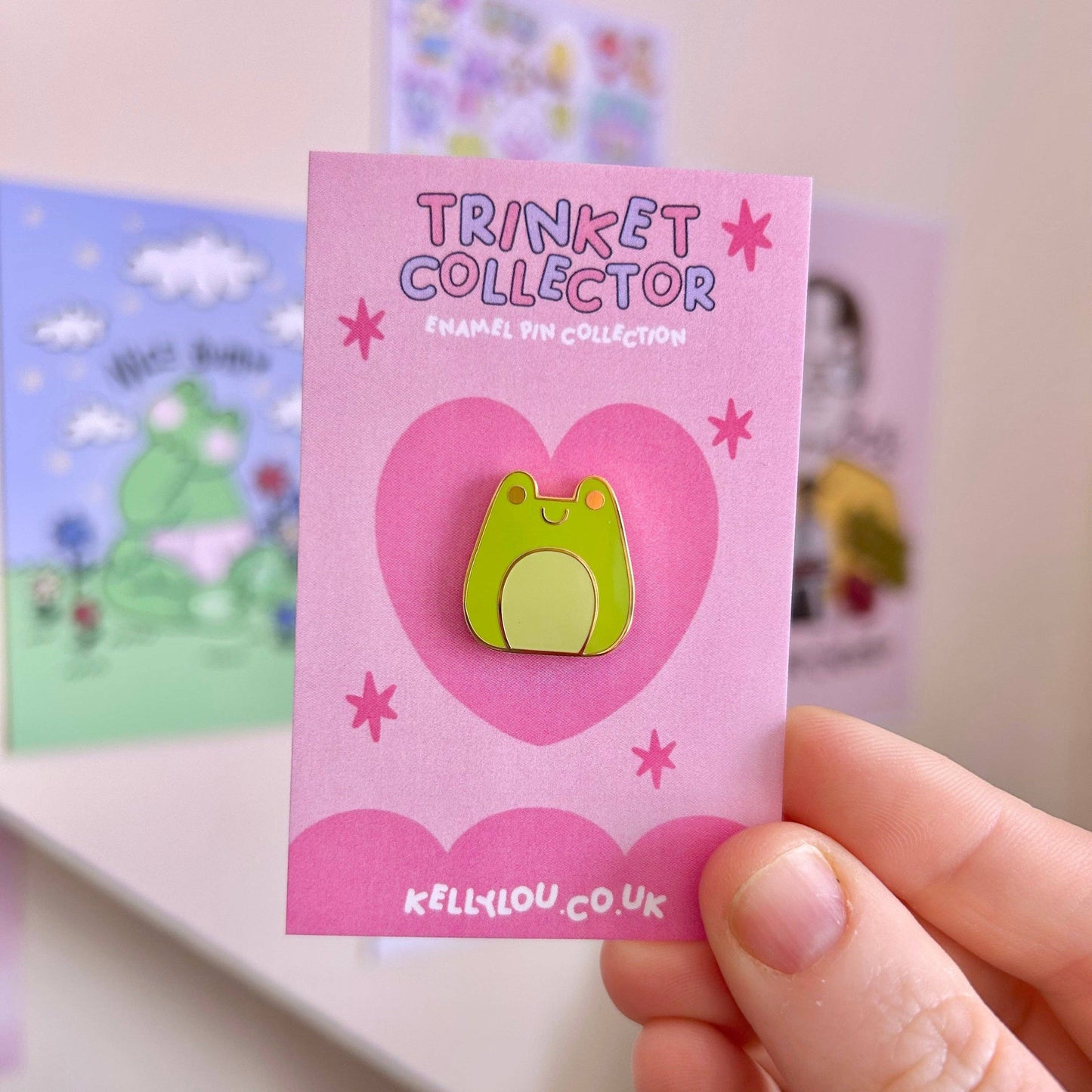Pink card with a green frog enamel pin and 'Trinket Collector' text, held by a hand.