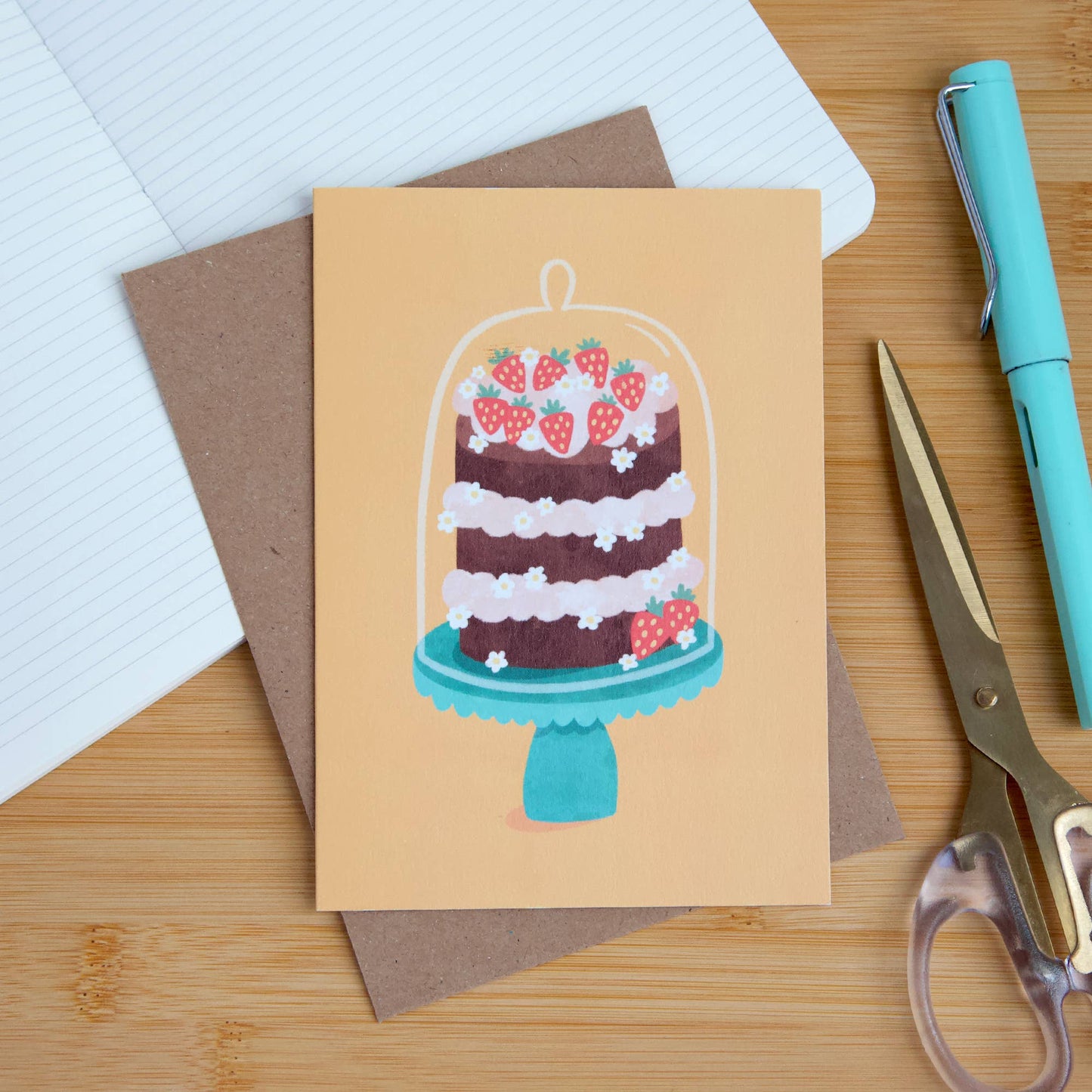 Greeting card with cake illustration on a wooden surface with stationery items.