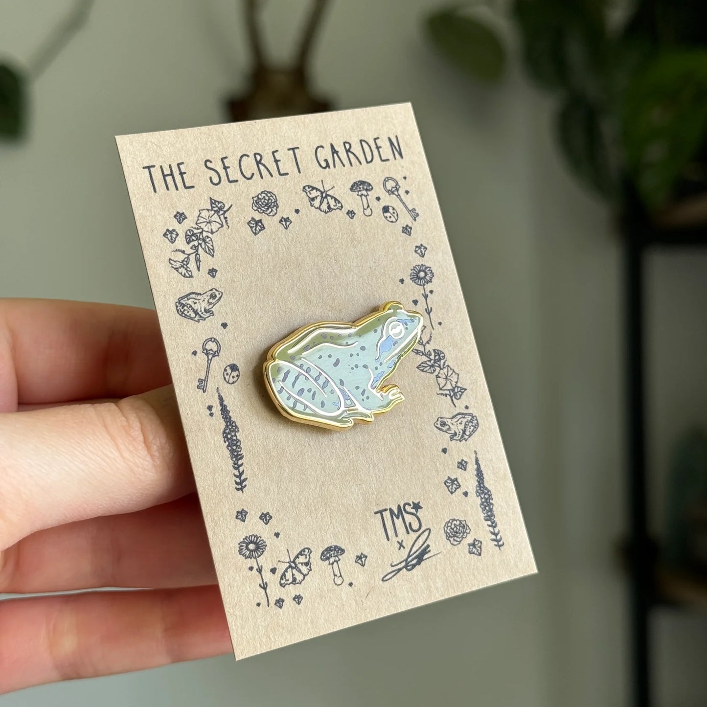 Frog-shaped pin on a 'The Secret Garden' card held by a hand