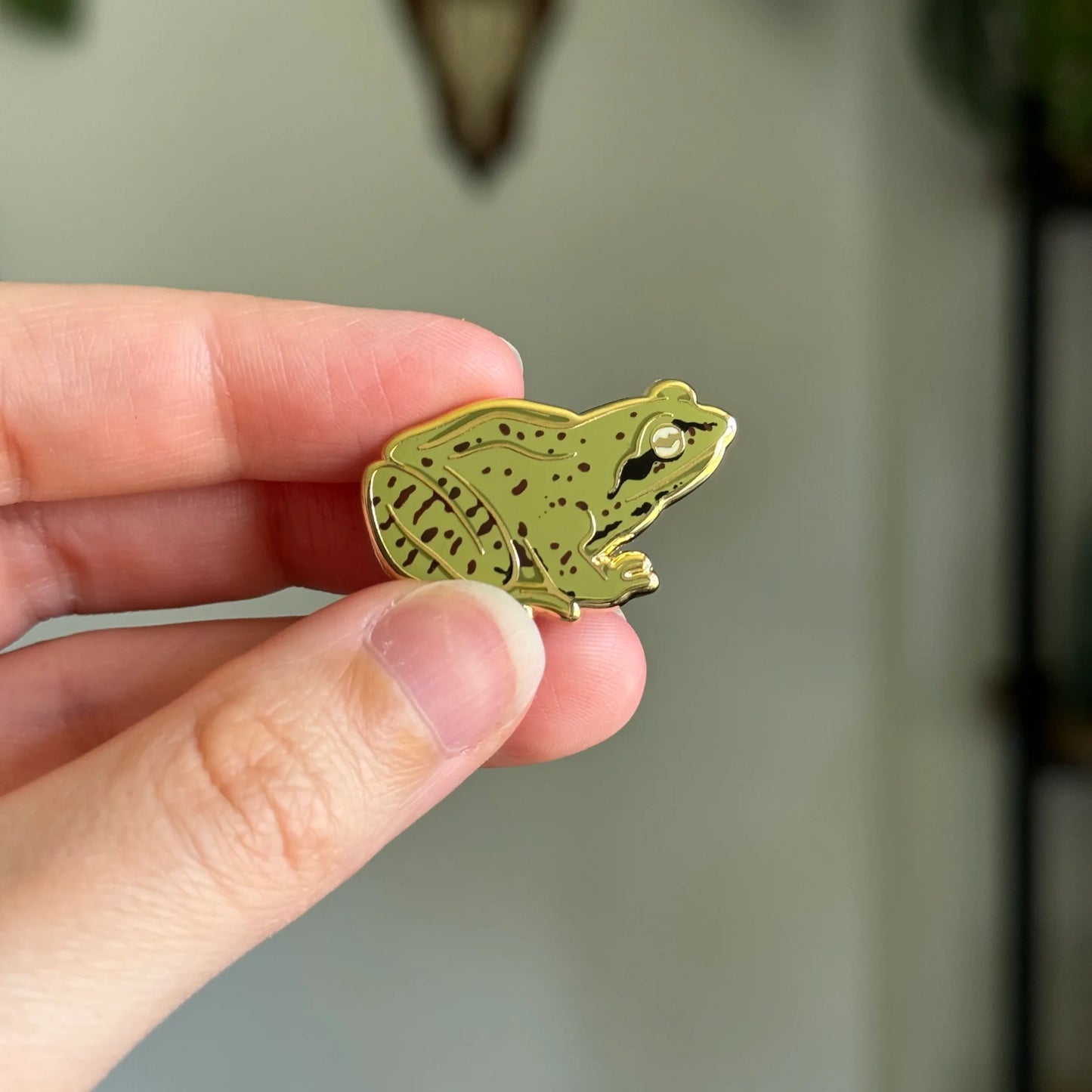 Gold frog-shaped pin held between fingers against a neutral background