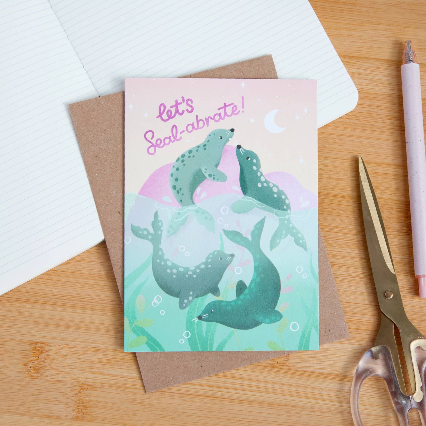 Greeting card with seals illustration and 'let's Seal-abrate!' text on a wooden surface with stationery items.