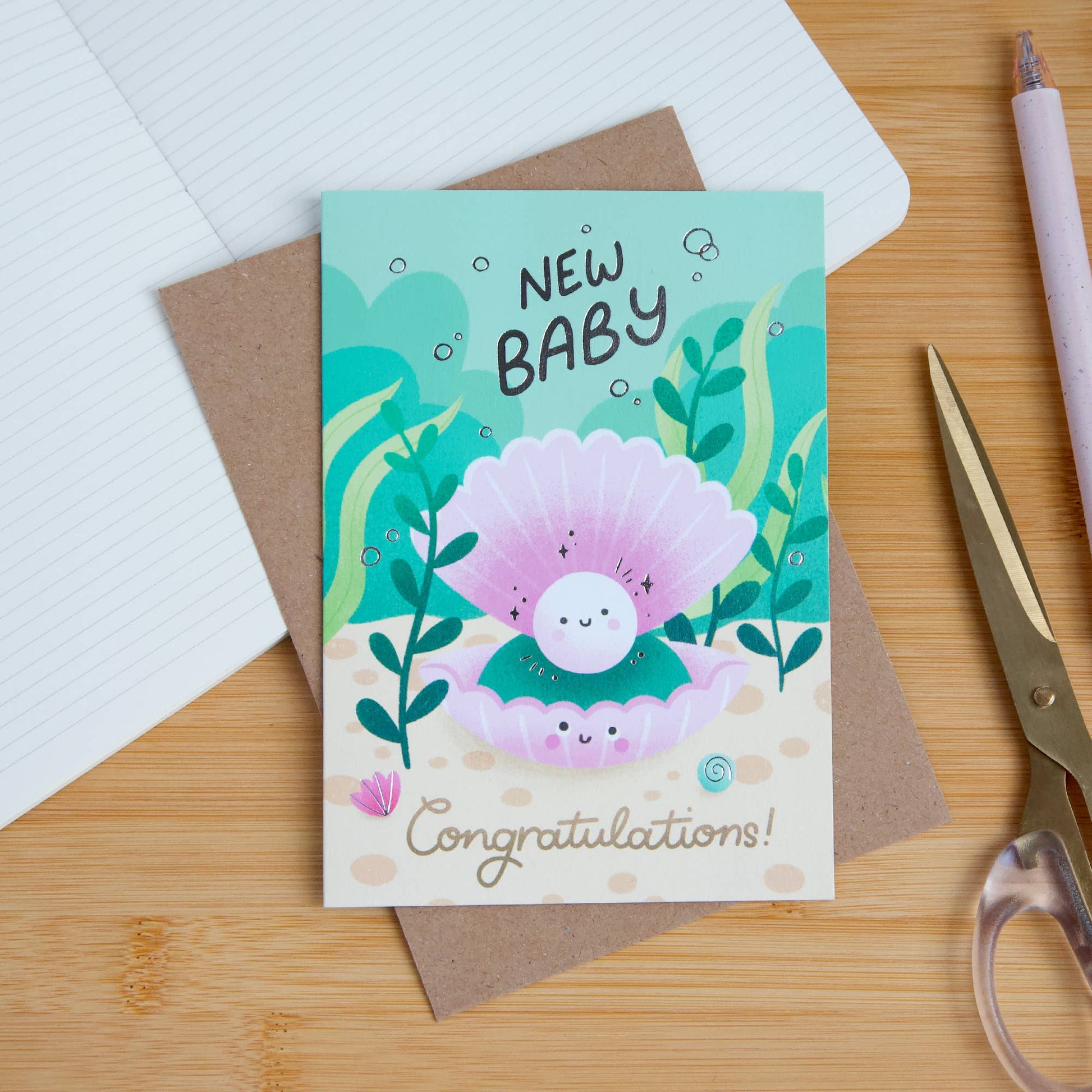 New baby card with a cartoon pearl and text on a wooden surface with stationery items.