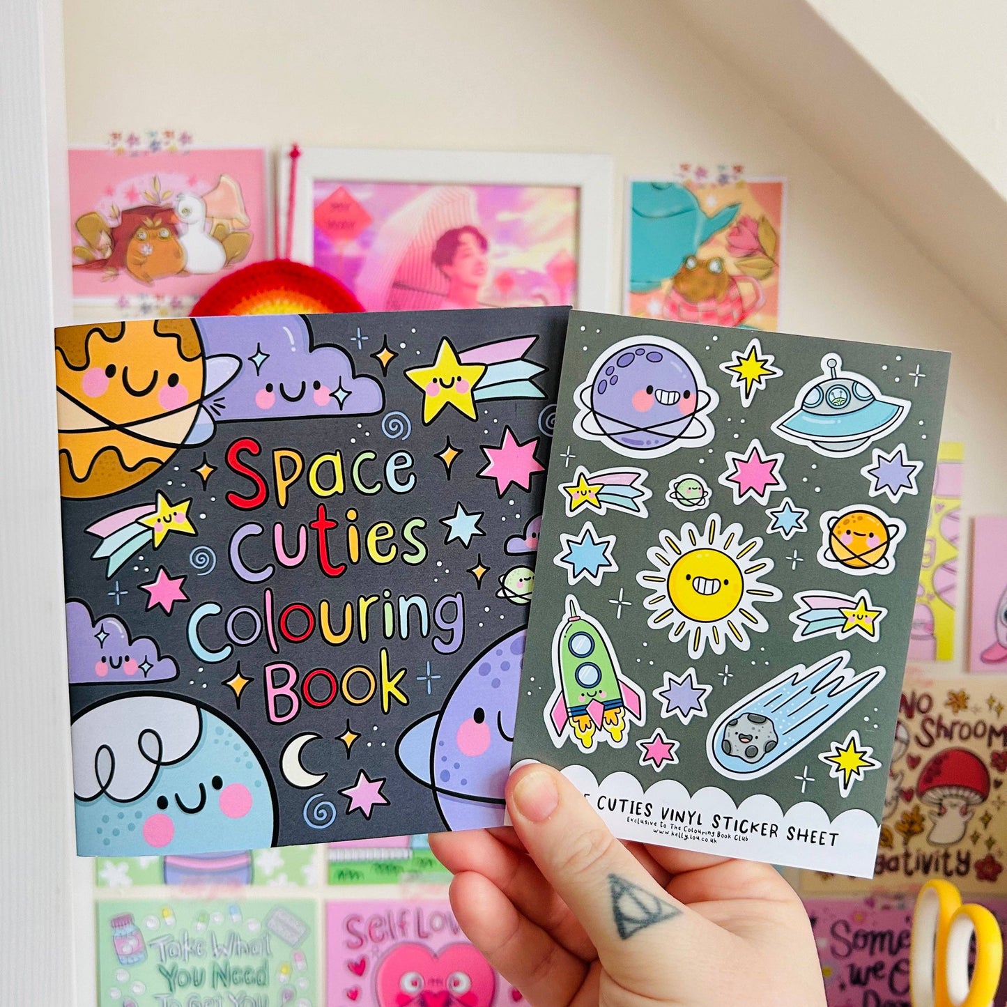 Two children's books titled 'Space Cuties Colouring Book' and 'Cuties Vinyl Sticker Sheet' held by a hand.
