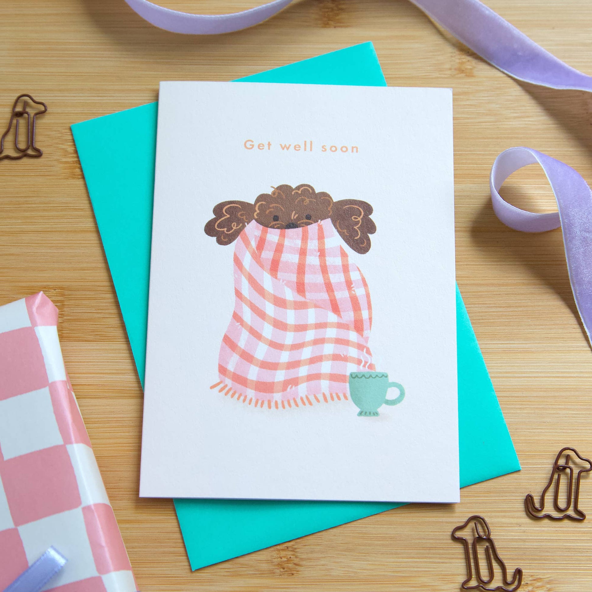 Greeting card with illustration of a person wrapped in a blanket and a cup, surrounded by stationery items on a wooden surface.