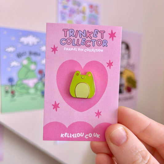 Pink card with a green frog enamel pin and 'Trinket Collector' text, held by a hand.