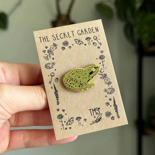 Green frog pin on a 'The Secret Garden' card held by a hand with a blurred background
