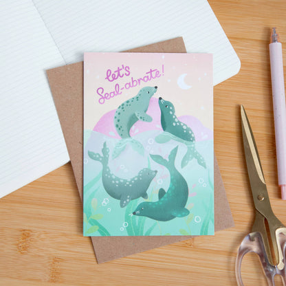 Greeting card with seals illustration and 'let's Seal-abrate!' text on a wooden surface with stationery items.