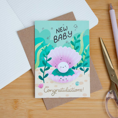 New baby card with a cartoon pearl and text on a wooden surface with stationery items.