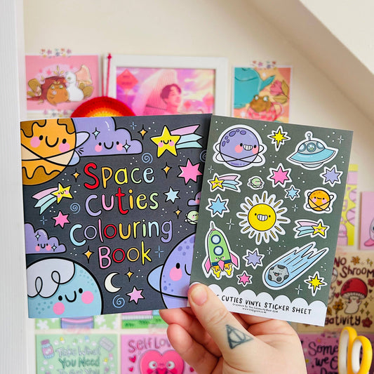 Two children's books titled 'Space Cuties Colouring Book' and 'Cuties Vinyl Sticker Sheet' held by a hand.