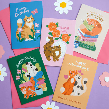 Collection of cat-themed greeting cards on a purple background with flowers.