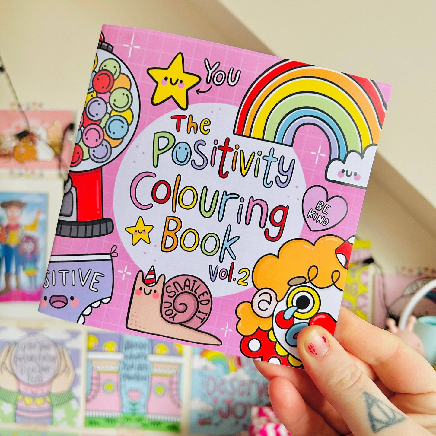 Positivity Colouring Book Vol. 2 held by a person with colorful illustrations on a pink background.