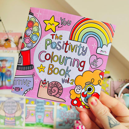 Positivity Colouring Book Vol. 2 held by a person with colorful illustrations on a pink background.
